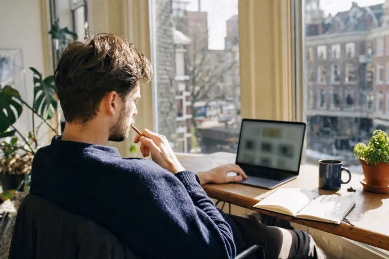 Webdesigner aan een houten bureau in een zonnig Amsterdams appartement, denkend met een potlood bij zijn laptop
