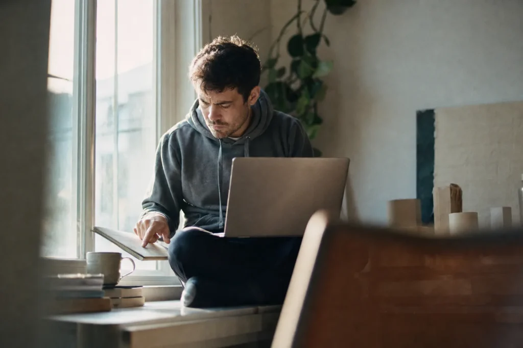 Developer op een vensterbank met laptop op schoot die een regel in zijn notitieboek volgt