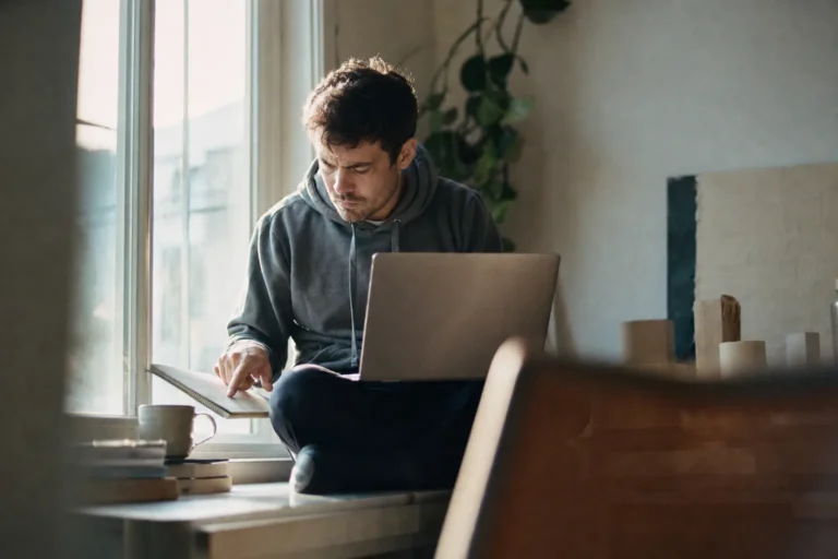 Developer op een vensterbank met laptop op schoot die een regel in zijn notitieboek volgt